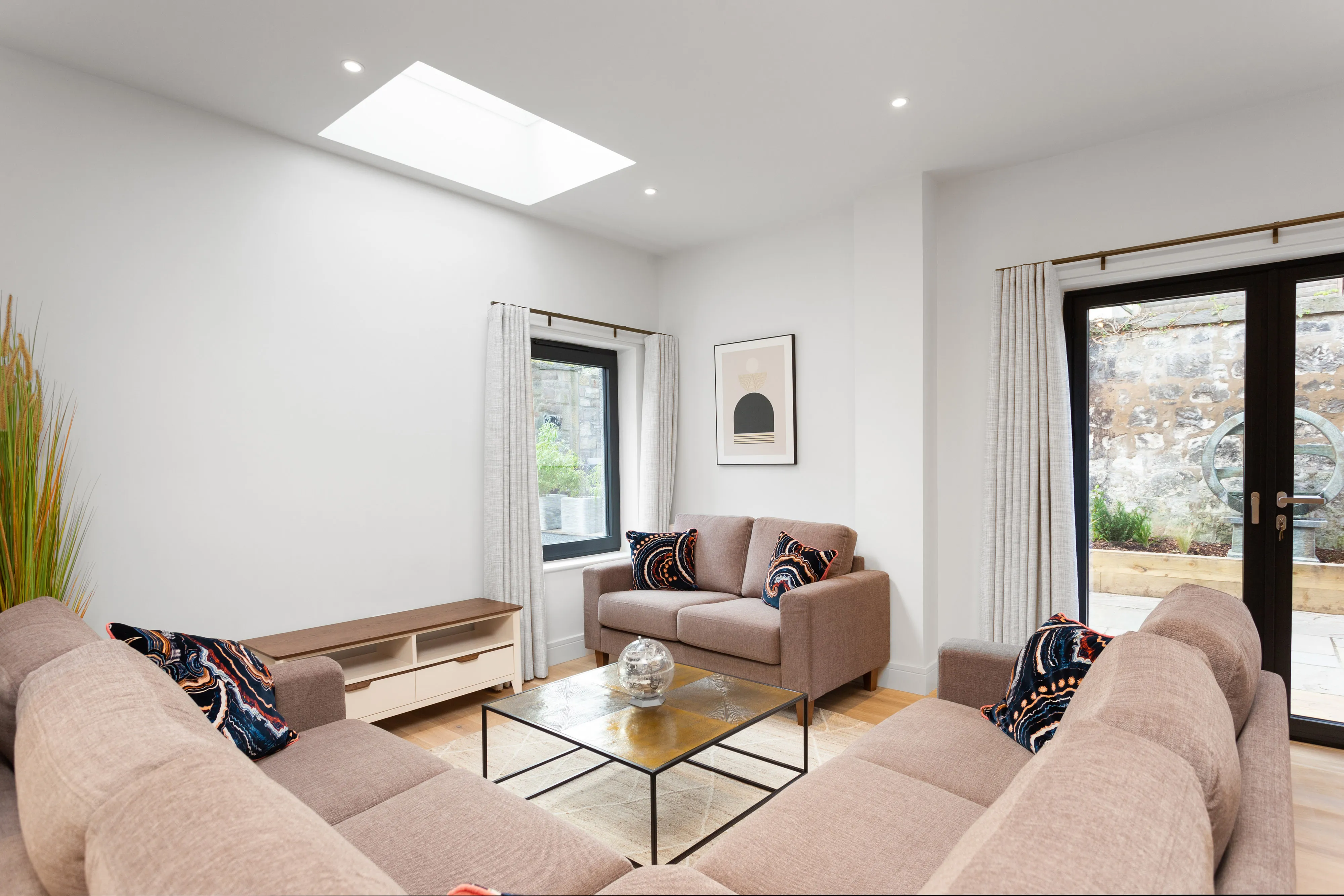 Living room with two blush sofas in patterned cushions around a glass coffee table, curtained windows either side, and glazed garden doors under a skylight.