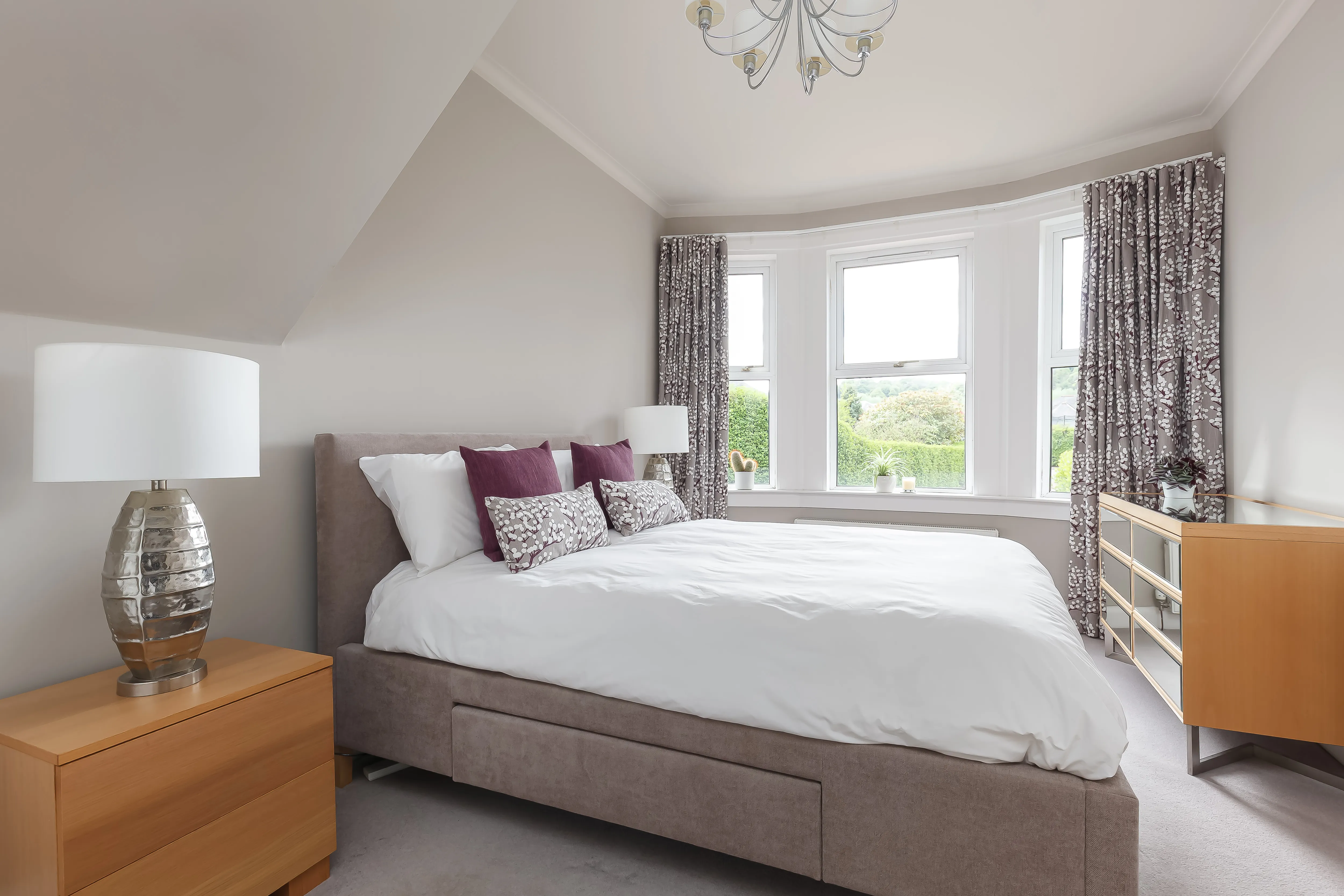 Bedroom bay window with full-height grey wave curtains in a blossom-branch print, either side of three sash windows behind an upholstered bed.