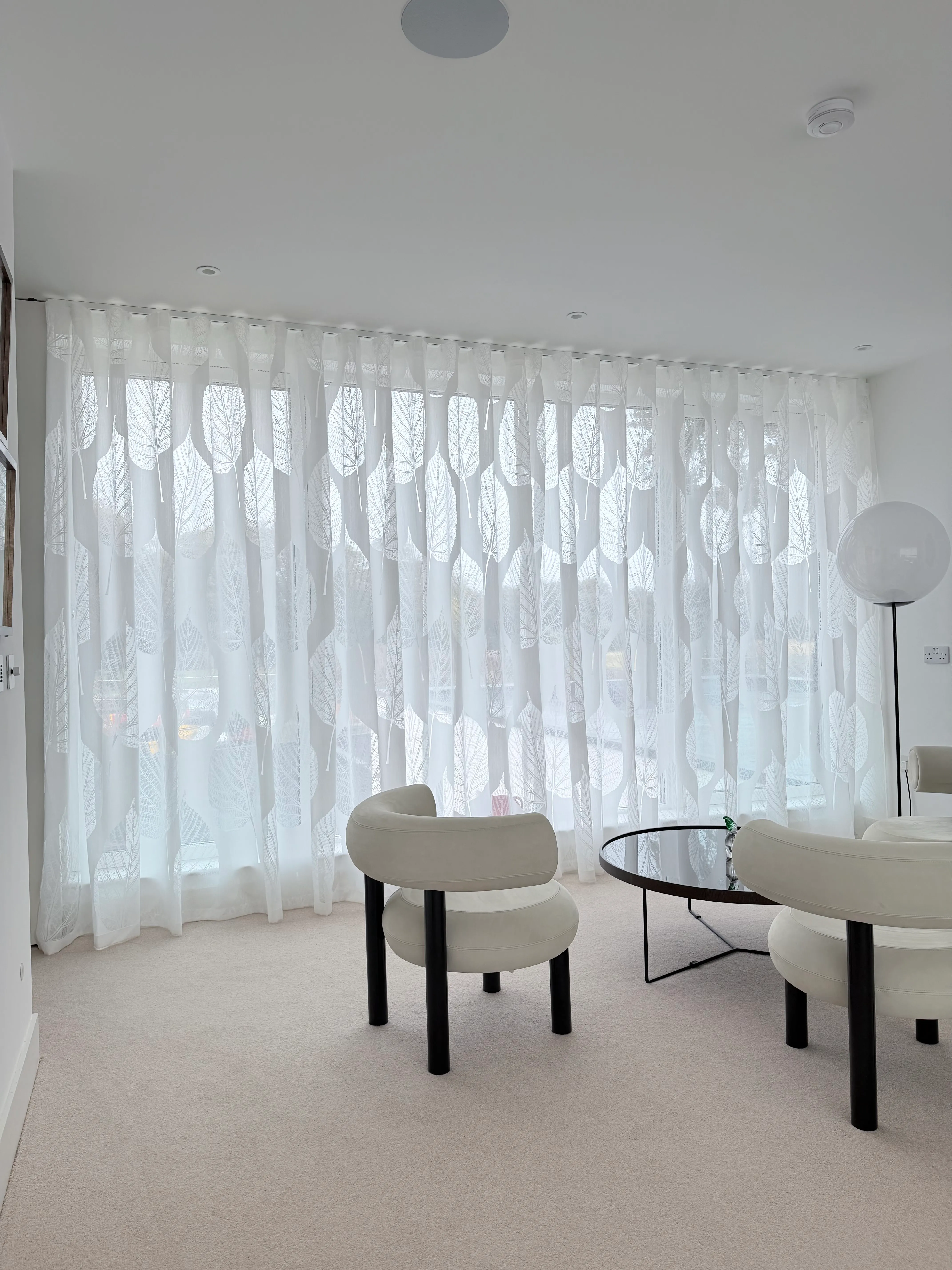 Floor-to-ceiling wave-headed sheer curtains in a leaf print across a wide window, with cream bouclé chairs and a dark round coffee table on beige carpet.