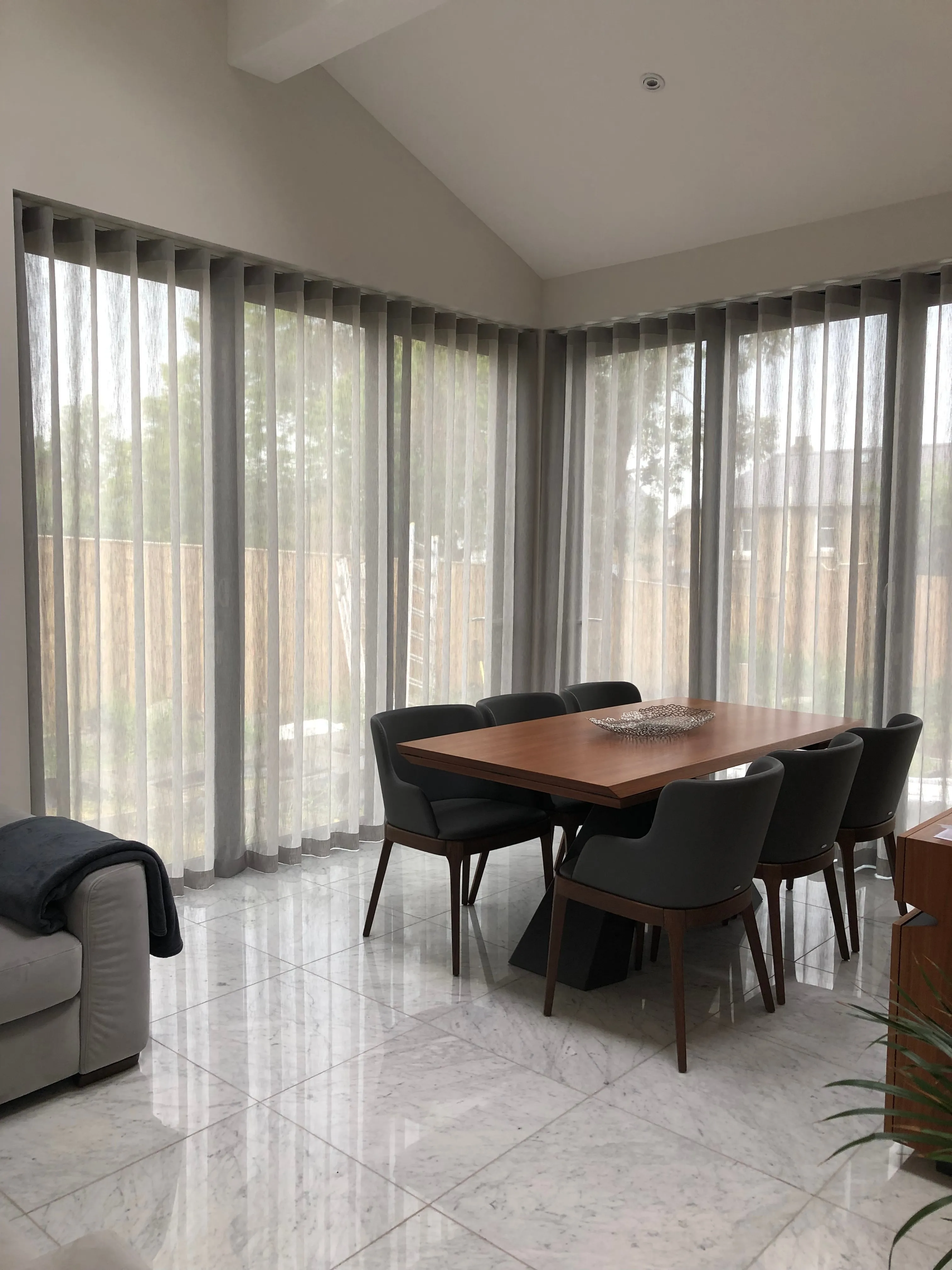 Vaulted dining room corner with full-height grey wave sheers wrapping two walls of glazing, a walnut table with charcoal upholstered chairs, and a marble-effect tiled floor.